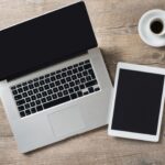 laptop and tablet on table with coffee, pen, cellphone, and eyeglasses.